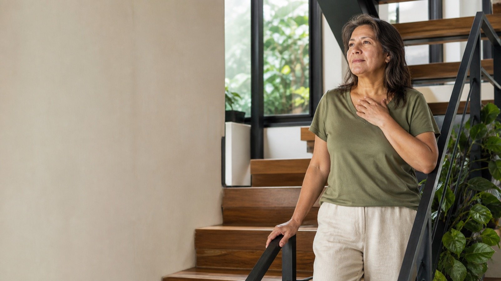 Mujer adulta haciendo una pausa consciente en casa, junto a una escalera y plantas.